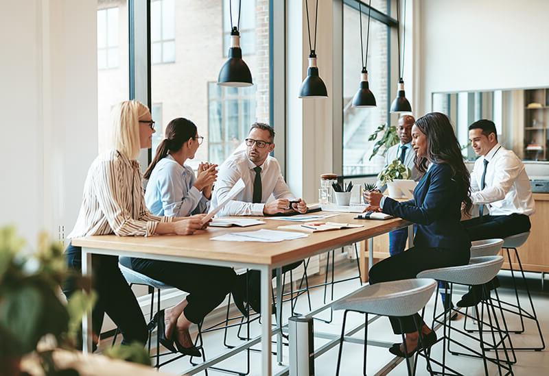 A group of six various people gather round a meeting room table in a modern office environment